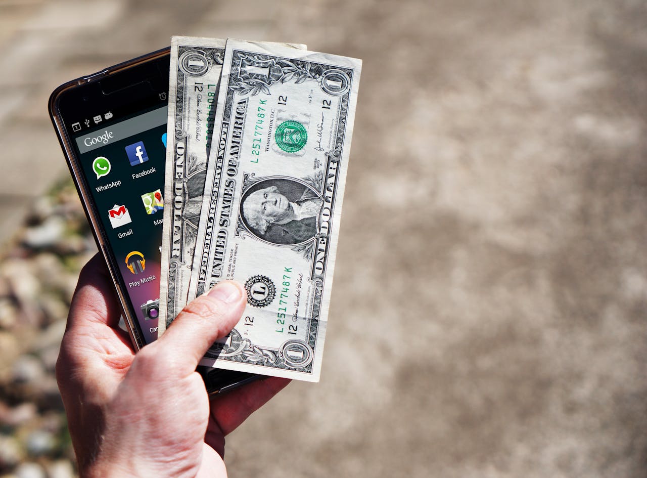 about-us Close-up of a hand holding US dollar bills and a smartphone outdoors, showcasing financial technology.