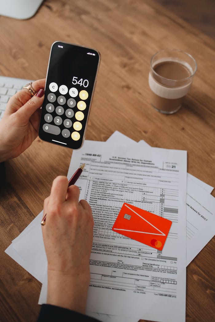 A person uses a smartphone calculator over tax documents on a wooden table.