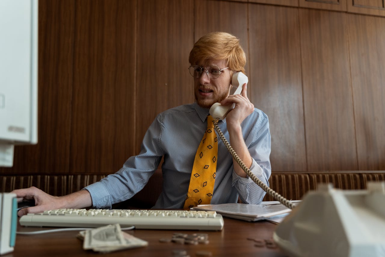 A man in business attire using a vintage telephone and computer in an office setting.